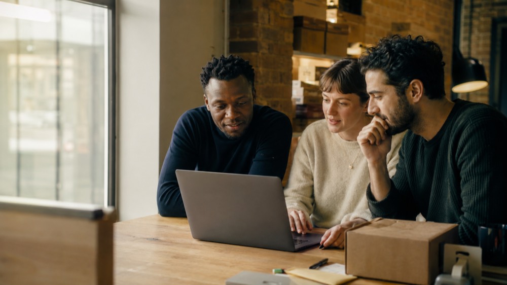Three colleagues leaning in around a laptop, working through something together at a shared desk.